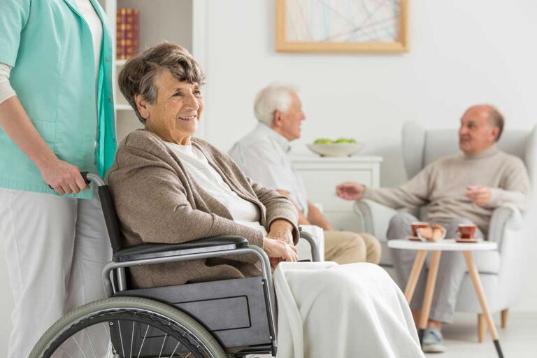 Older adults and a caregiver share a calm conversation in a bright assisted living common room.